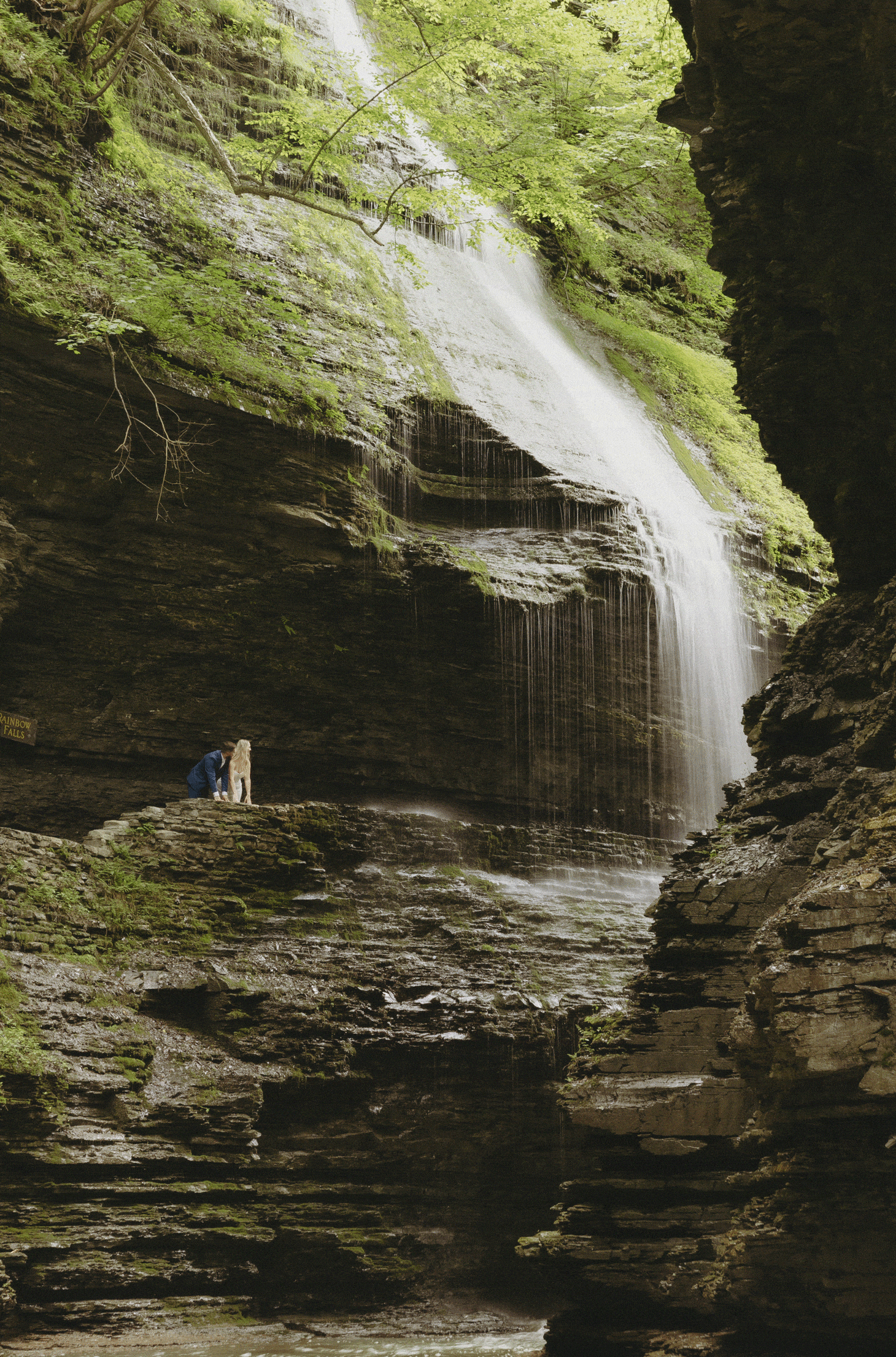 A couple in wedding attire stands by a stone wall in a rocky canyon with a waterfall and lush greenery in the background for Watkins Glen State Park Wedding Photos