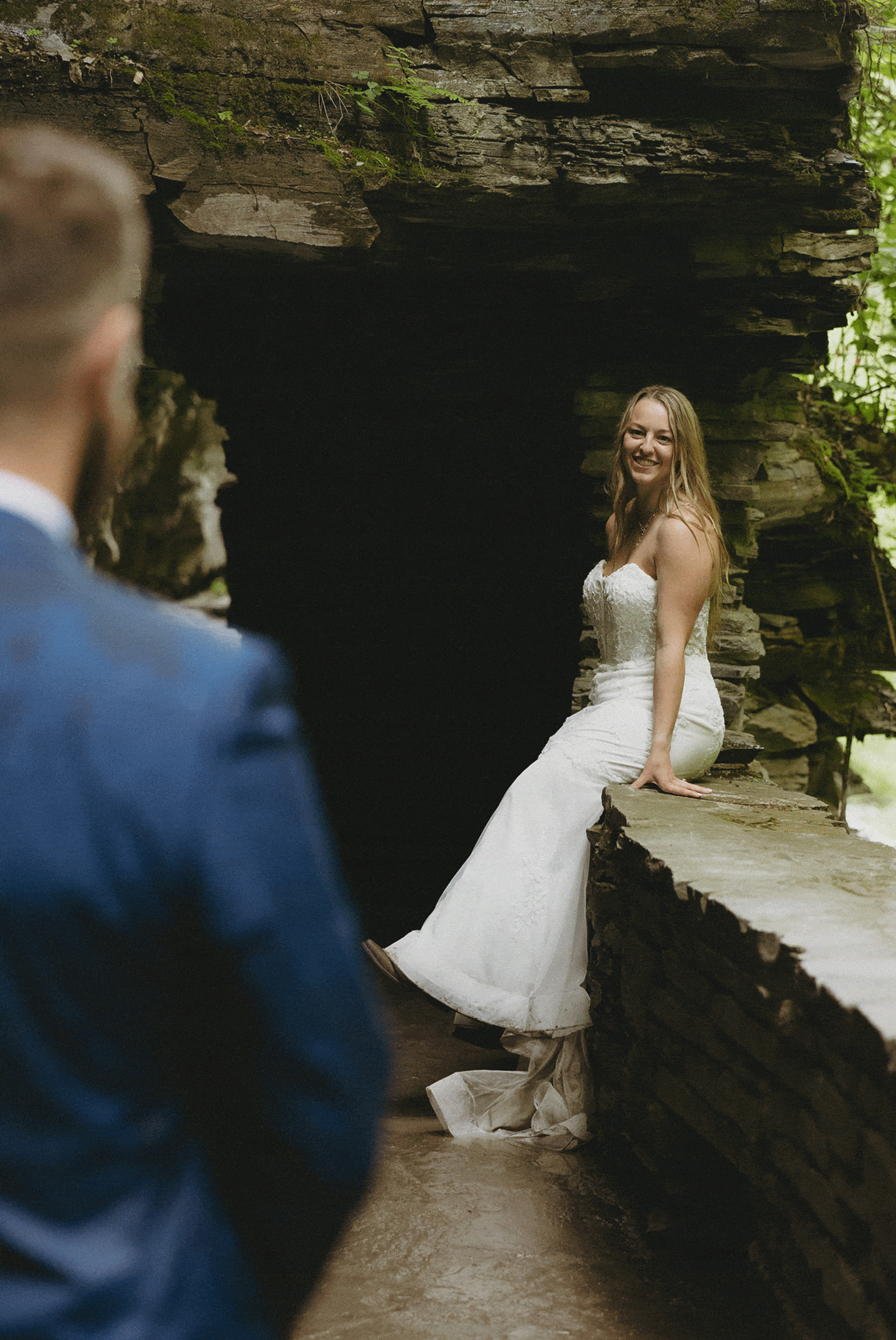 A bride in a white dress sits on a stone ledge, smiling, while a man in a blue suit stands in the foreground, partially out of focus.