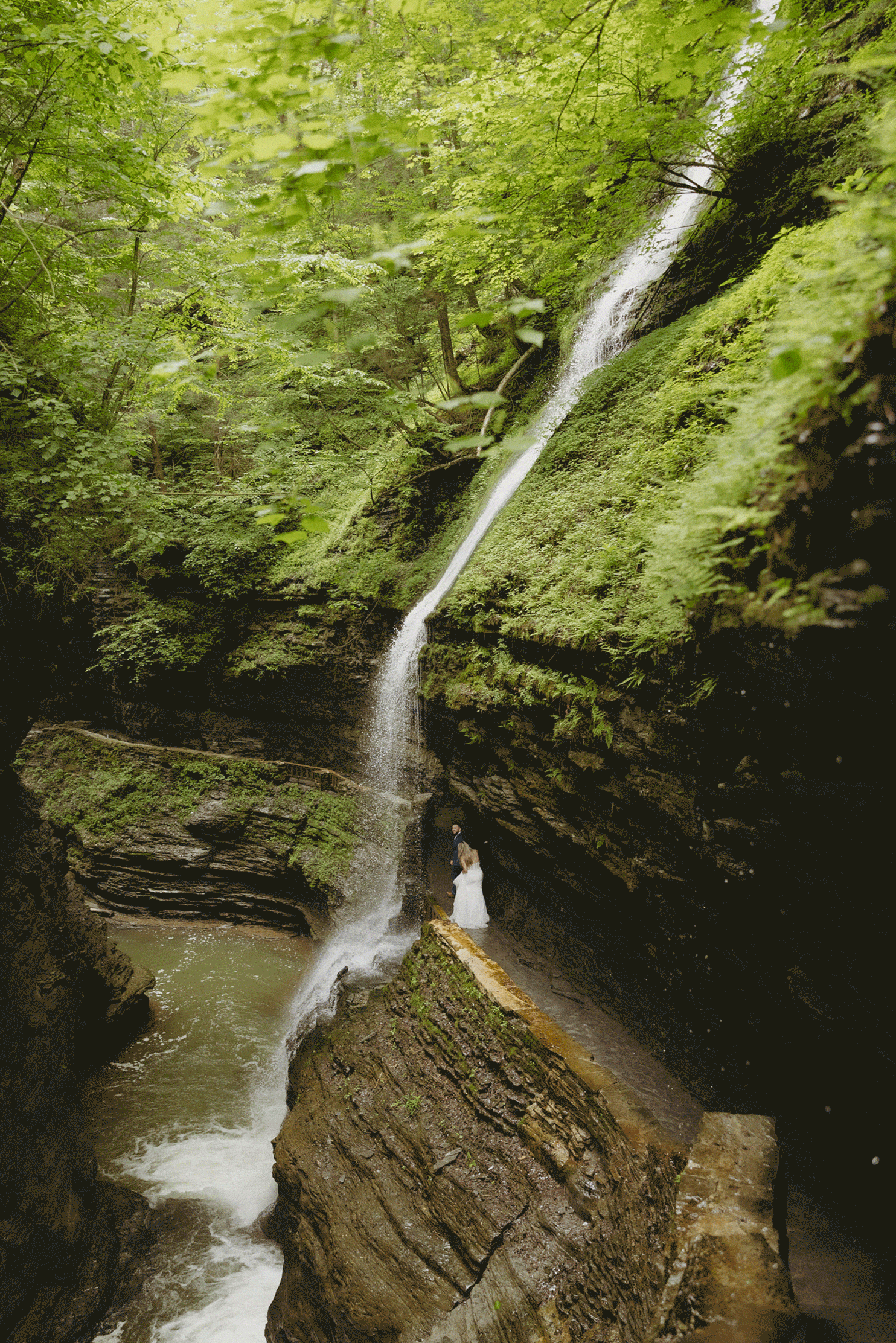 A person in white stands near a tall waterfall cascading down mossy rocks, surrounded by lush green foliage in a forested area.
