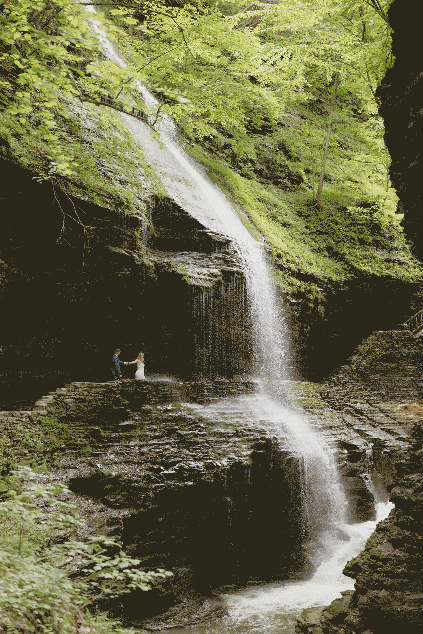 A person in white stands near a tall waterfall cascading down mossy rocks, surrounded by lush green foliage in a forested area.