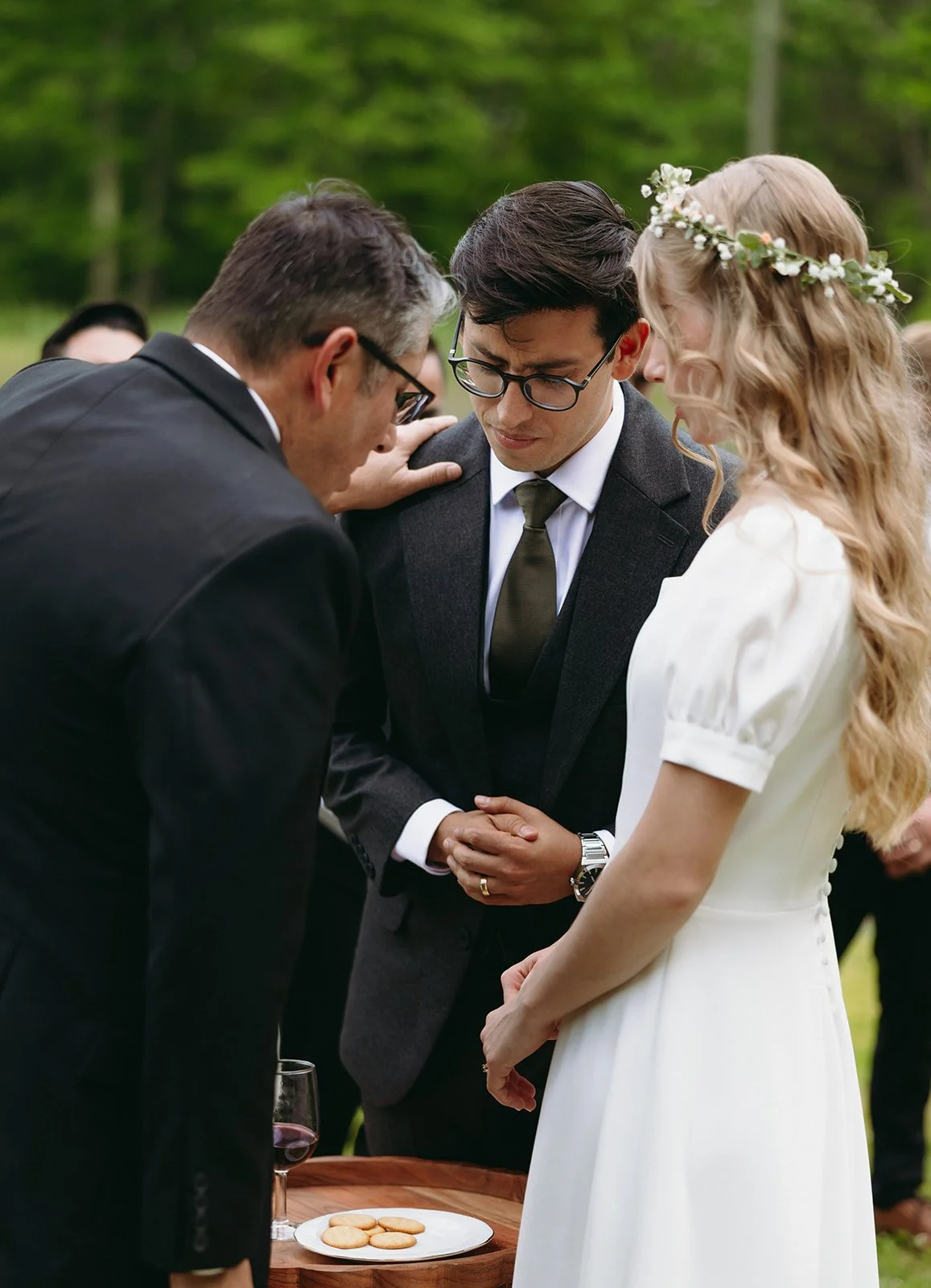 A man and woman stand with heads bowed and hands clasped, in front of a man in a suit, outdoors. | Christian wedding ceremony ideas