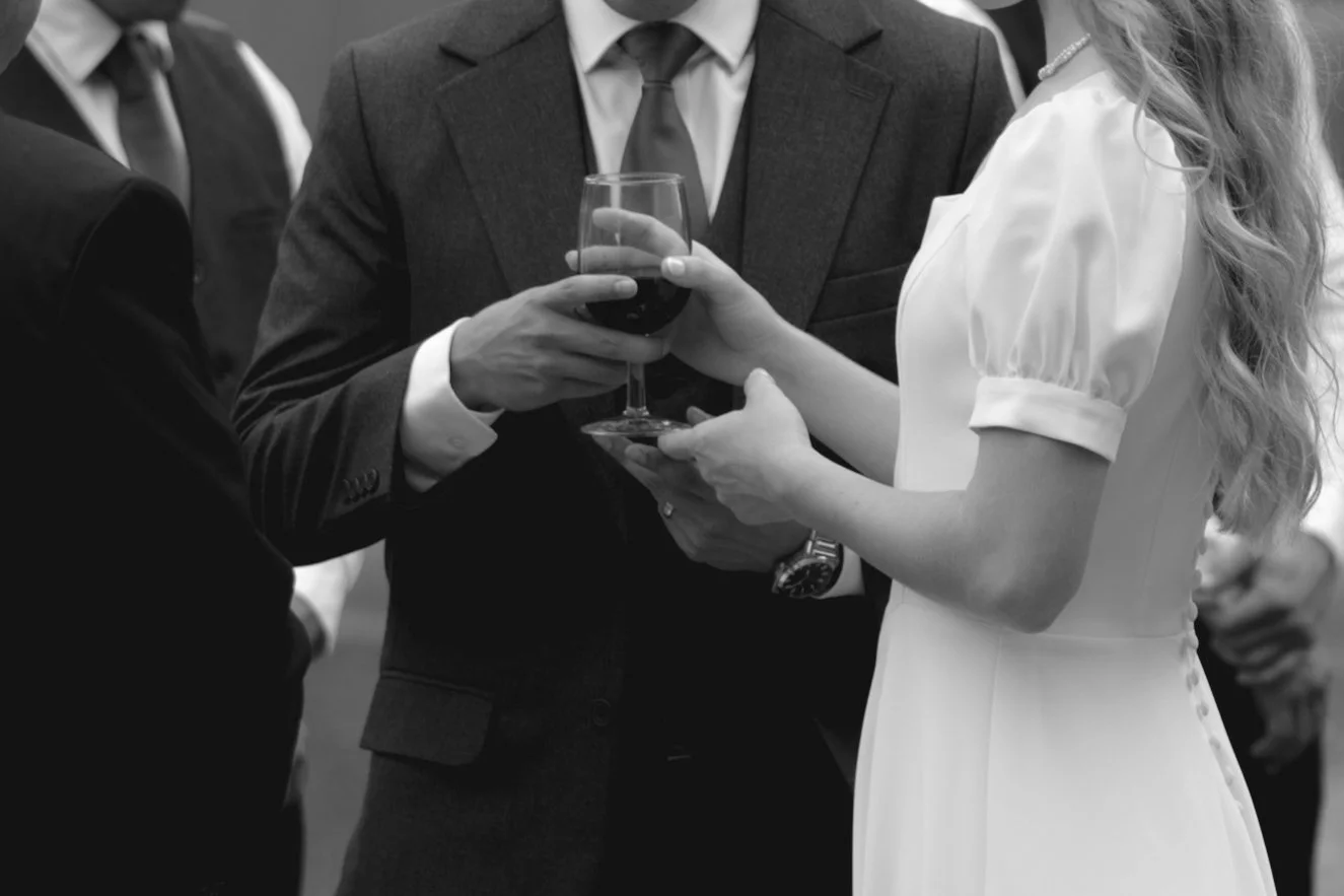 A man in a suit holds a wine glass while a woman in a white dress stands next to him at a formal event.