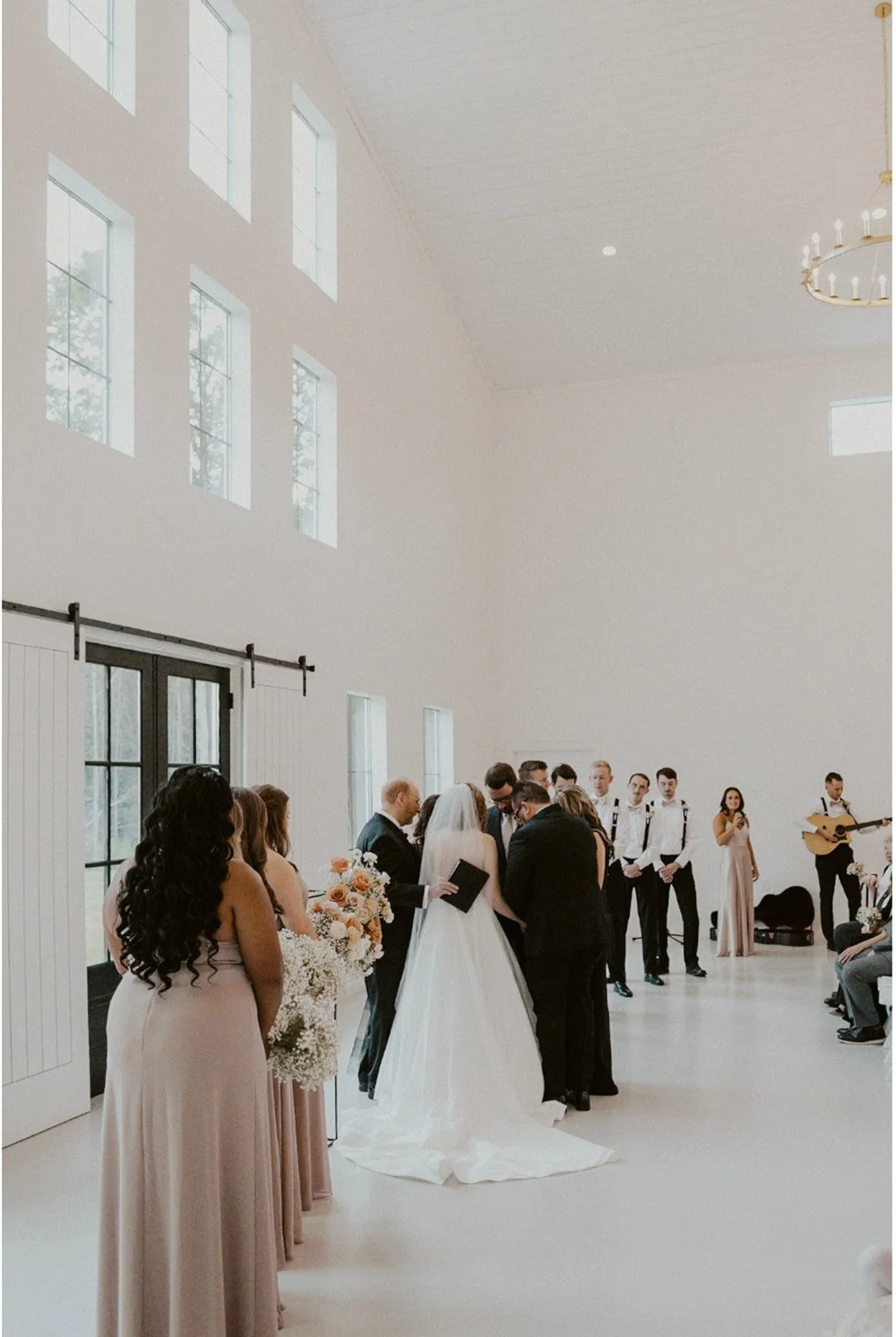 A bride and groom stand together at the altar during a wedding ceremony inside a bright, modern venue with guests and bridal party present.