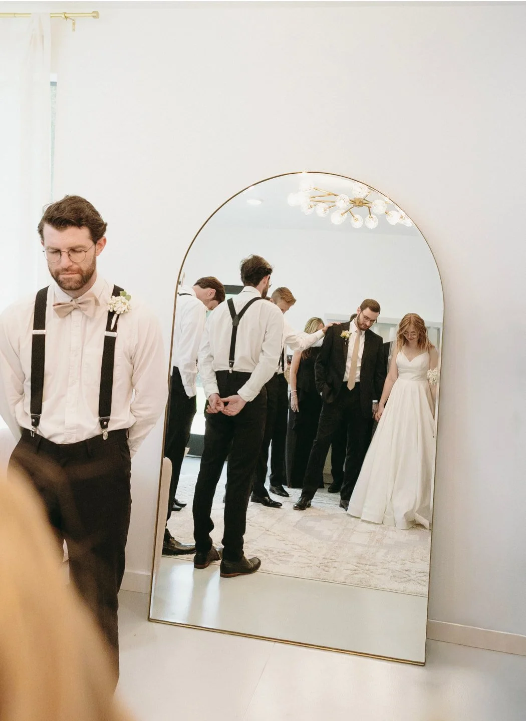 A man in suspenders stands in front of a mirror reflecting a bride, groom, and wedding party inside a bright, white room. | Christian wedding ceremony ideas 