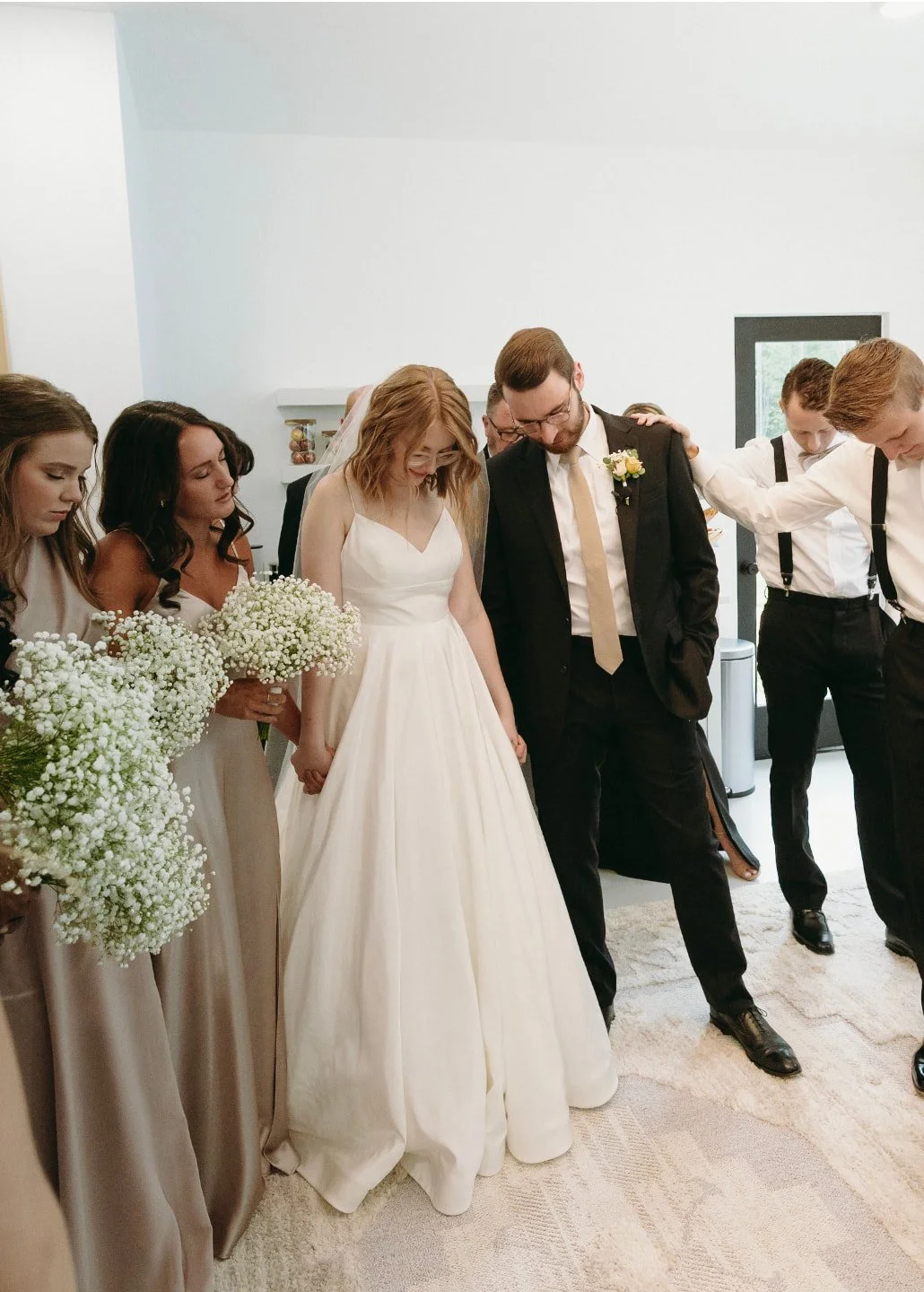 A man in suspenders stands in front of a mirror reflecting a bride, groom, and wedding party inside a bright, white room. | Christian wedding ceremony ideas 