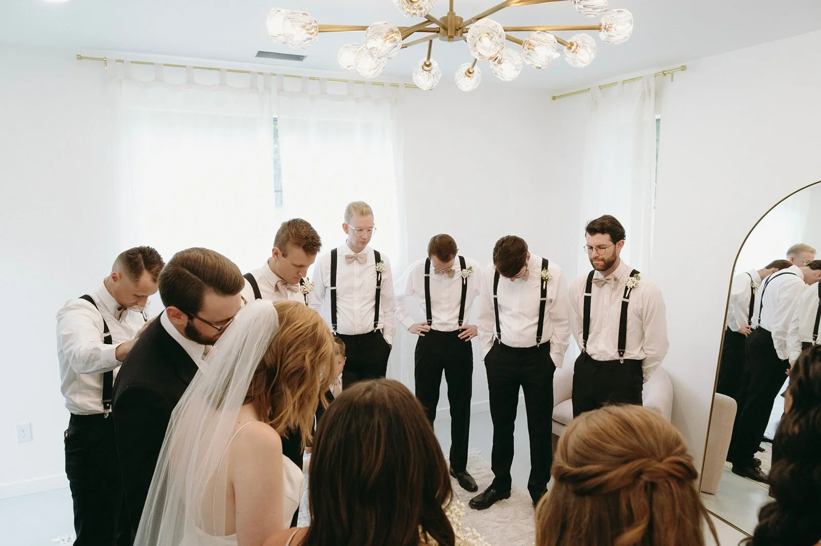 A man in suspenders stands in front of a mirror reflecting a bride, groom, and wedding party inside a bright, white room. | Christian wedding ceremony ideas 