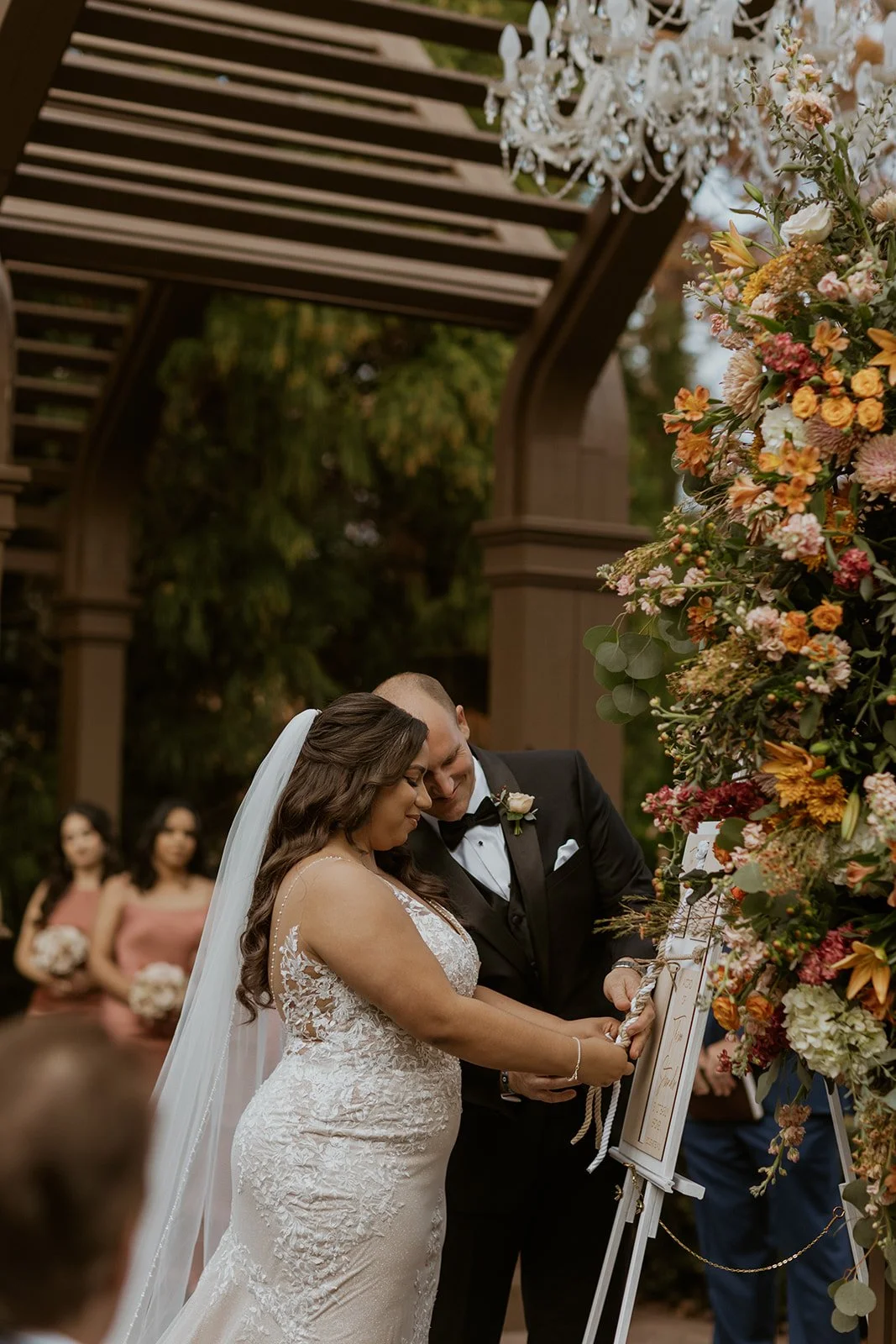 A bride and groom in formal attire stand together during an outdoor wedding ceremony, signing a document near a large floral arrangement. Bridesmaids holding bouquets stand in the background.