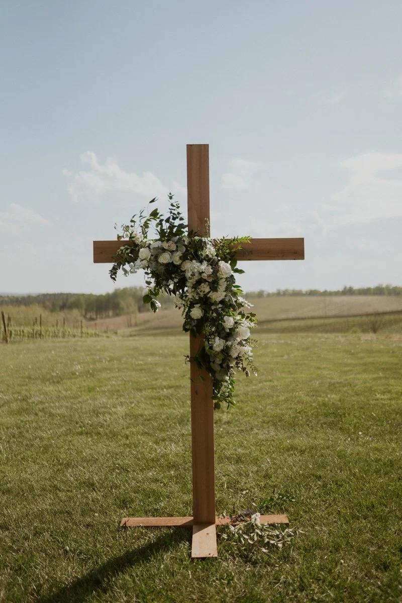 Wooden cross decorated with white flowers and greenery stands on a grassy field under a clear sky. | Christian wedding ceremony ideas