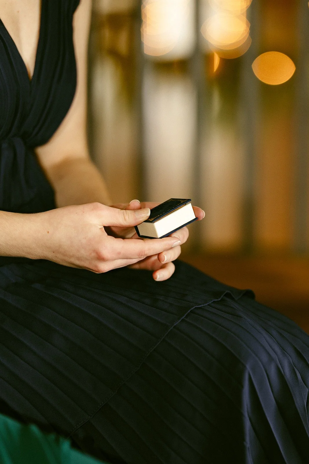 Person in a dark dress sitting and holding a small jewelry box with both hands; background is softly blurred with warm lighting.