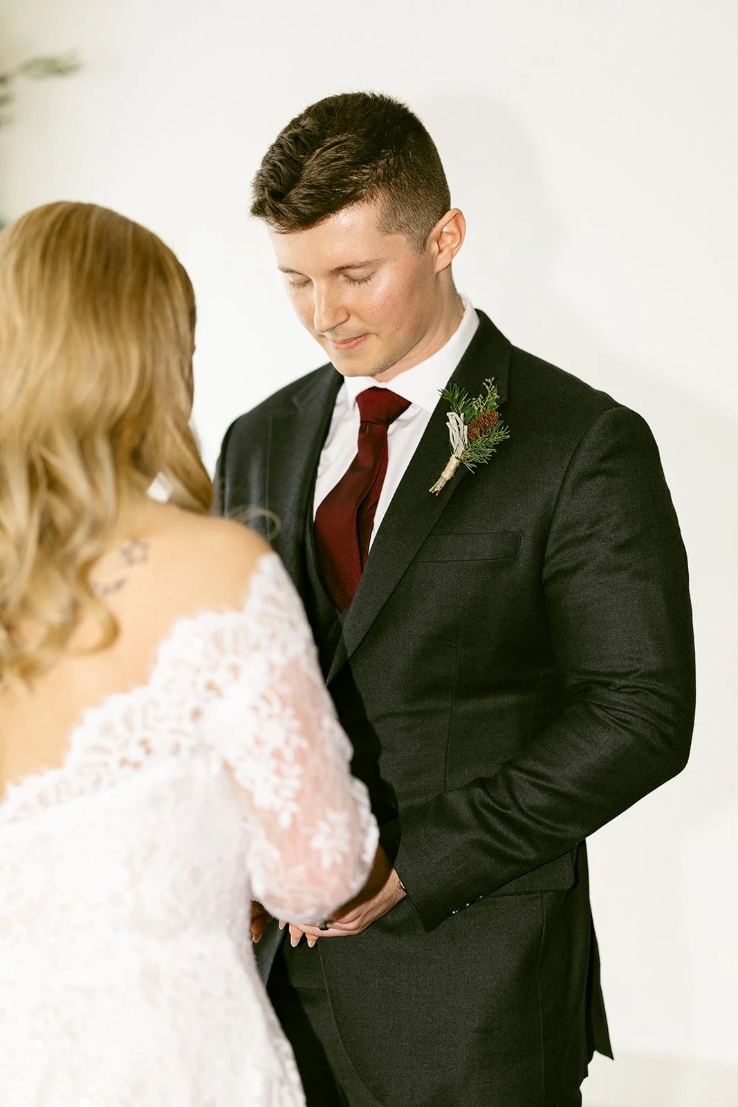 A bride and groom stand facing each other, holding hands during a wedding ceremony. The bride wears a white lace dress; the groom wears a dark suit with a maroon tie and boutonniere.