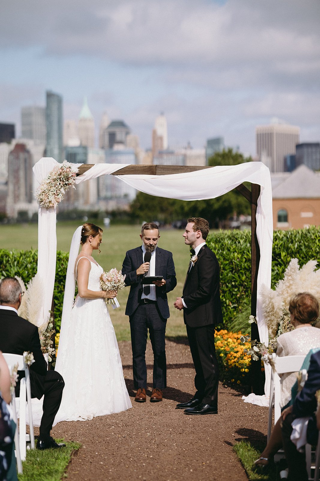 A bride and groom stand under a wedding arch outdoors with an officiant, exchanging vows for a christian wedding ceremony | Christian wedding ceremony ieas
