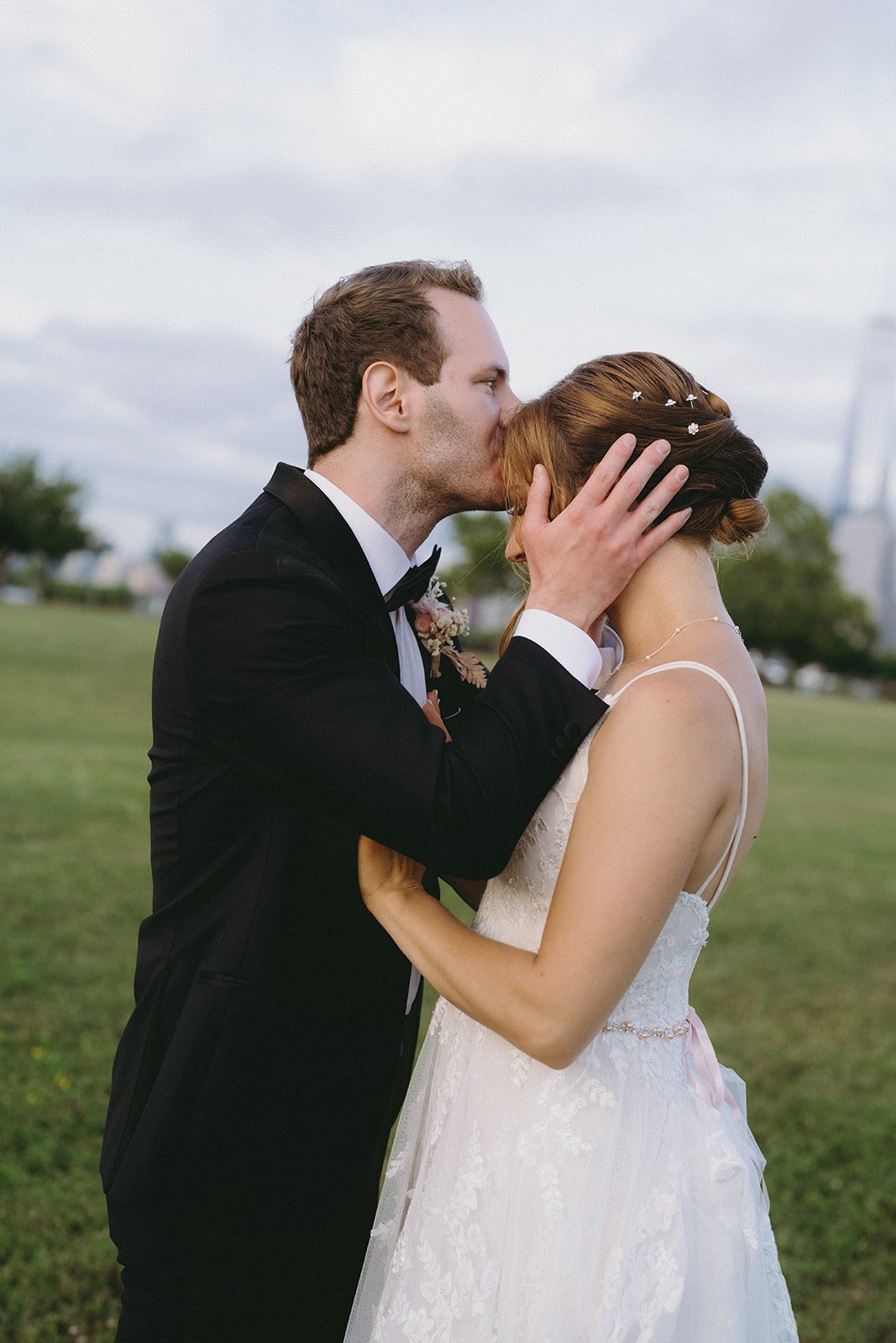 A groom in a black suit kisses the forehead of a bride in a white wedding dress as they stand together outdoors on a grassy field.