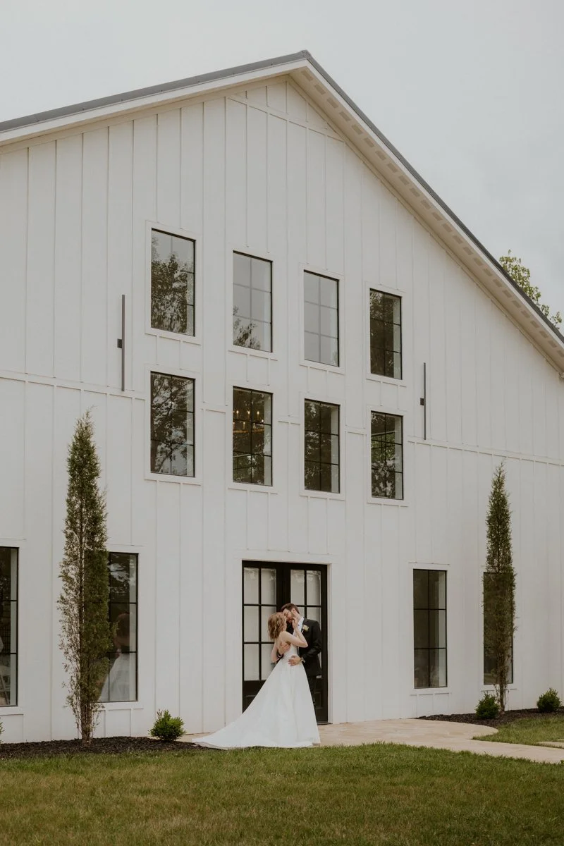 A bride and groom stand together outside a modern white barn with tall windows, posing for a wedding photo on a cloudy day.