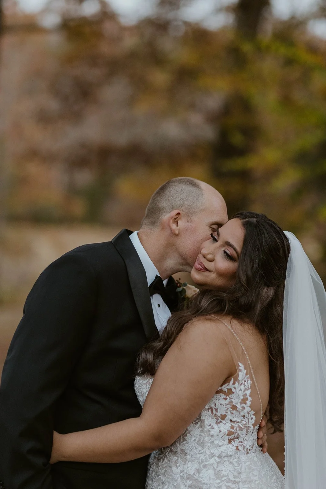 A groom in a black tuxedo kisses his bride, who wears a white lace wedding dress and veil, outdoors with blurred trees in the background.