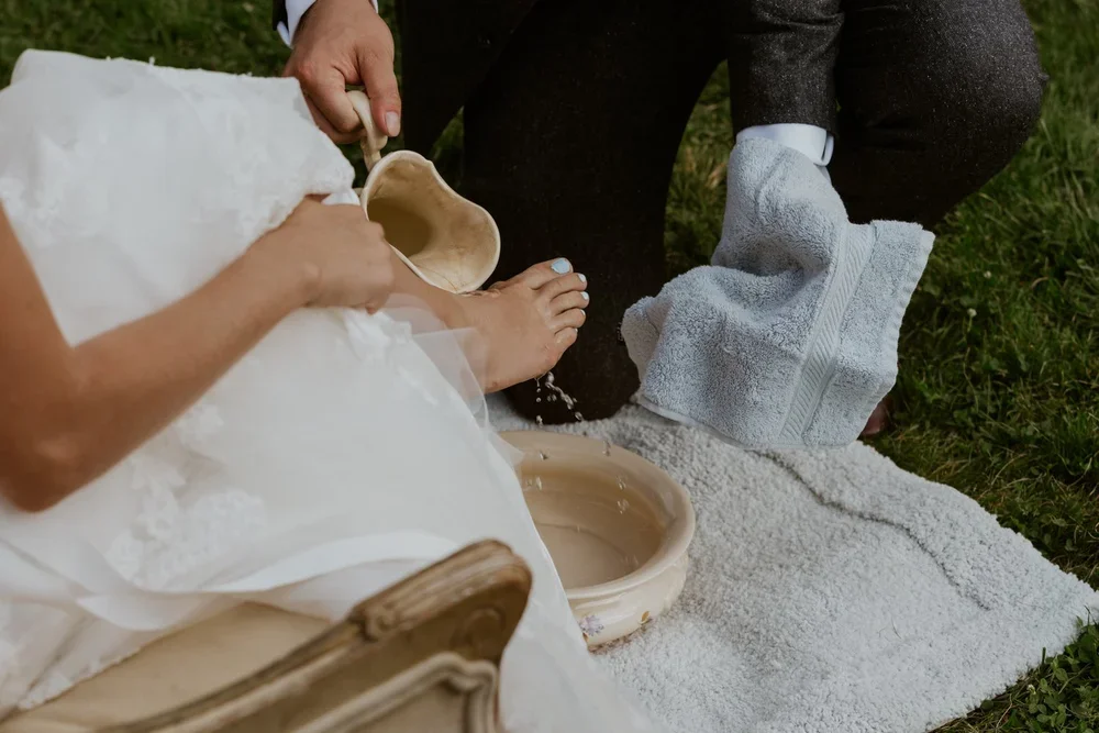 A person in a white dress has their foot washed by another person using a pitcher and bowl on a towel outdoors.