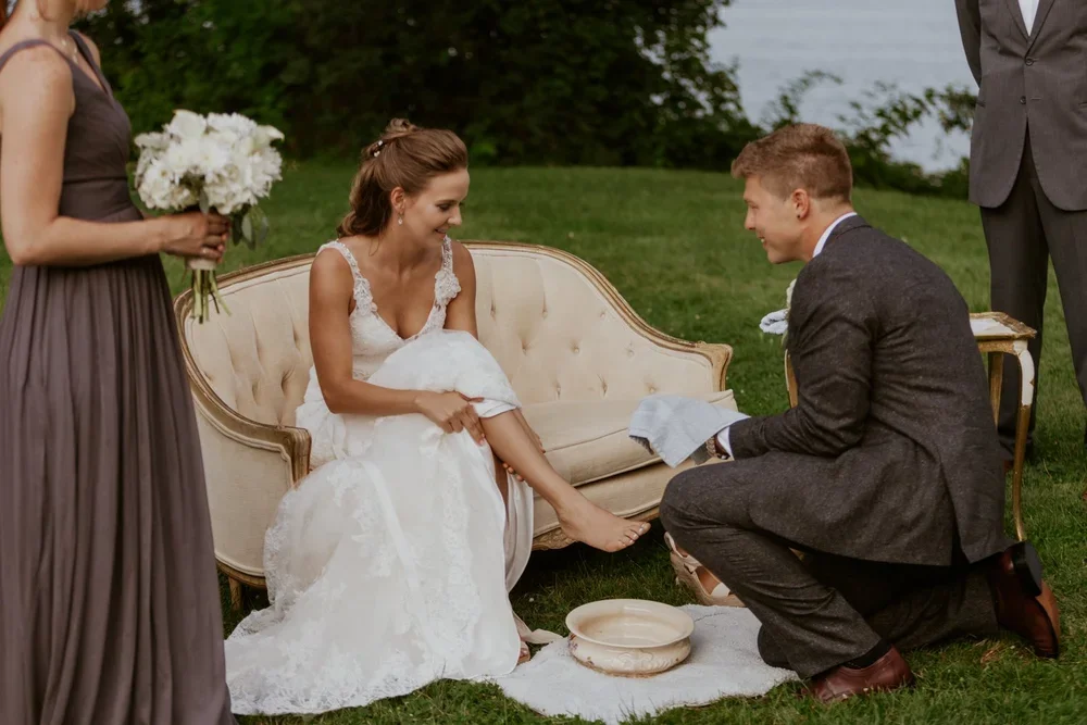A person in a white dress has their foot washed by another person using a pitcher and bowl on a towel outdoors.