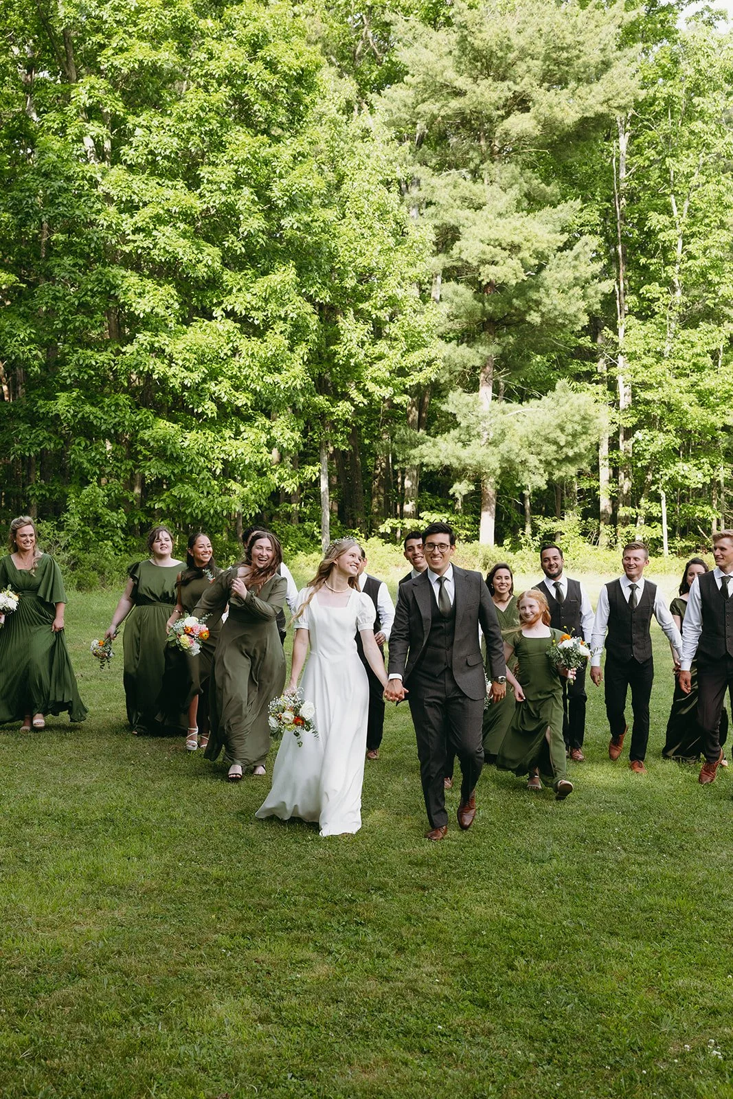 A bride and groom walk hand in hand outdoors, followed by their wedding party dressed in green and black attire, surrounded by trees and grass for christ centered marriage ceremony 