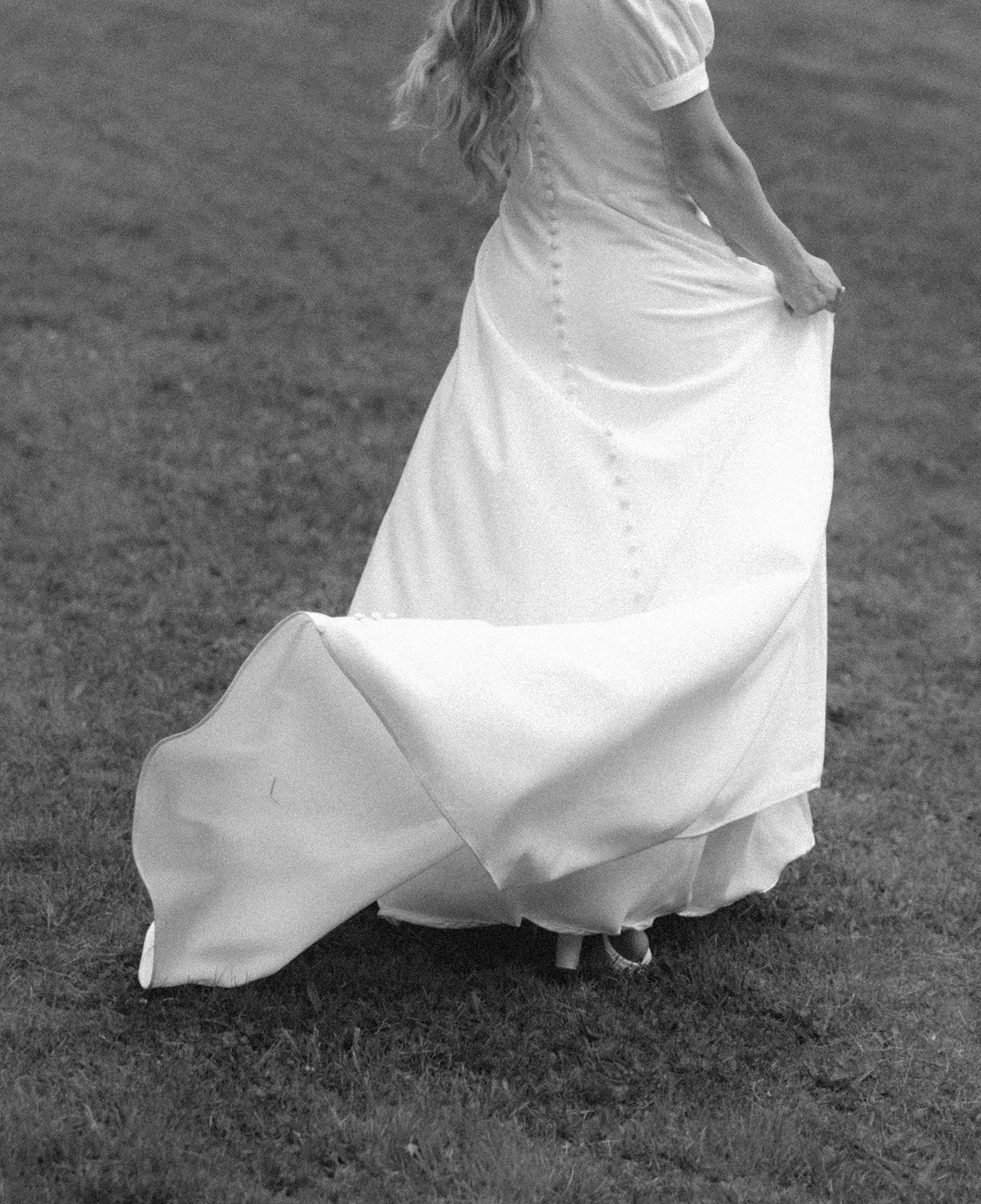 A person in a long, buttoned white dress walks on grass, holding up the skirt with one hand.