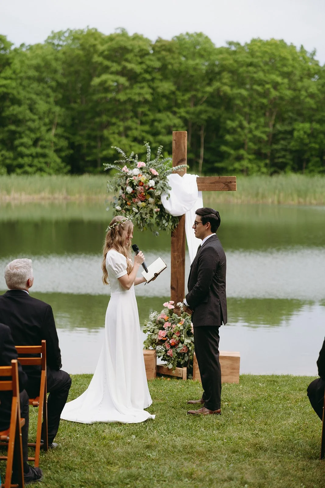 A bride and groom stand facing each other by a lake, reading vows in front of a wooden cross decorated with flowers, during an outdoor wedding ceremony for a christ centered marriage