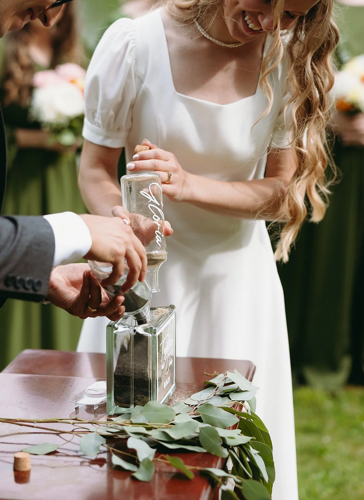 A bride and groom pour sand into a glass container during a wedding ceremony, with greenery on a wooden table in front of them