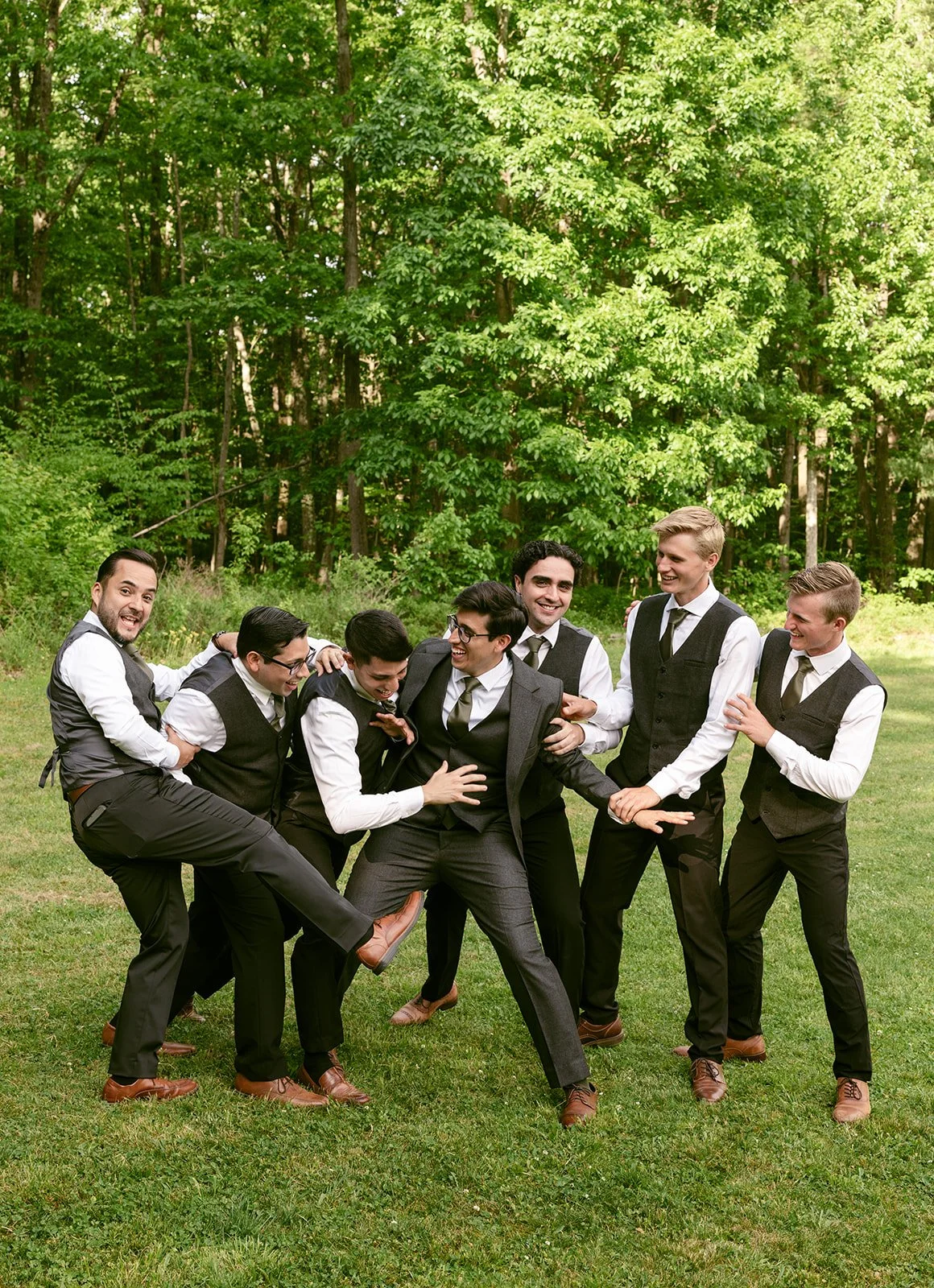Seven men in dress shirts, vests, and ties laugh and playfully jostle each other while standing on grass with trees in the background.