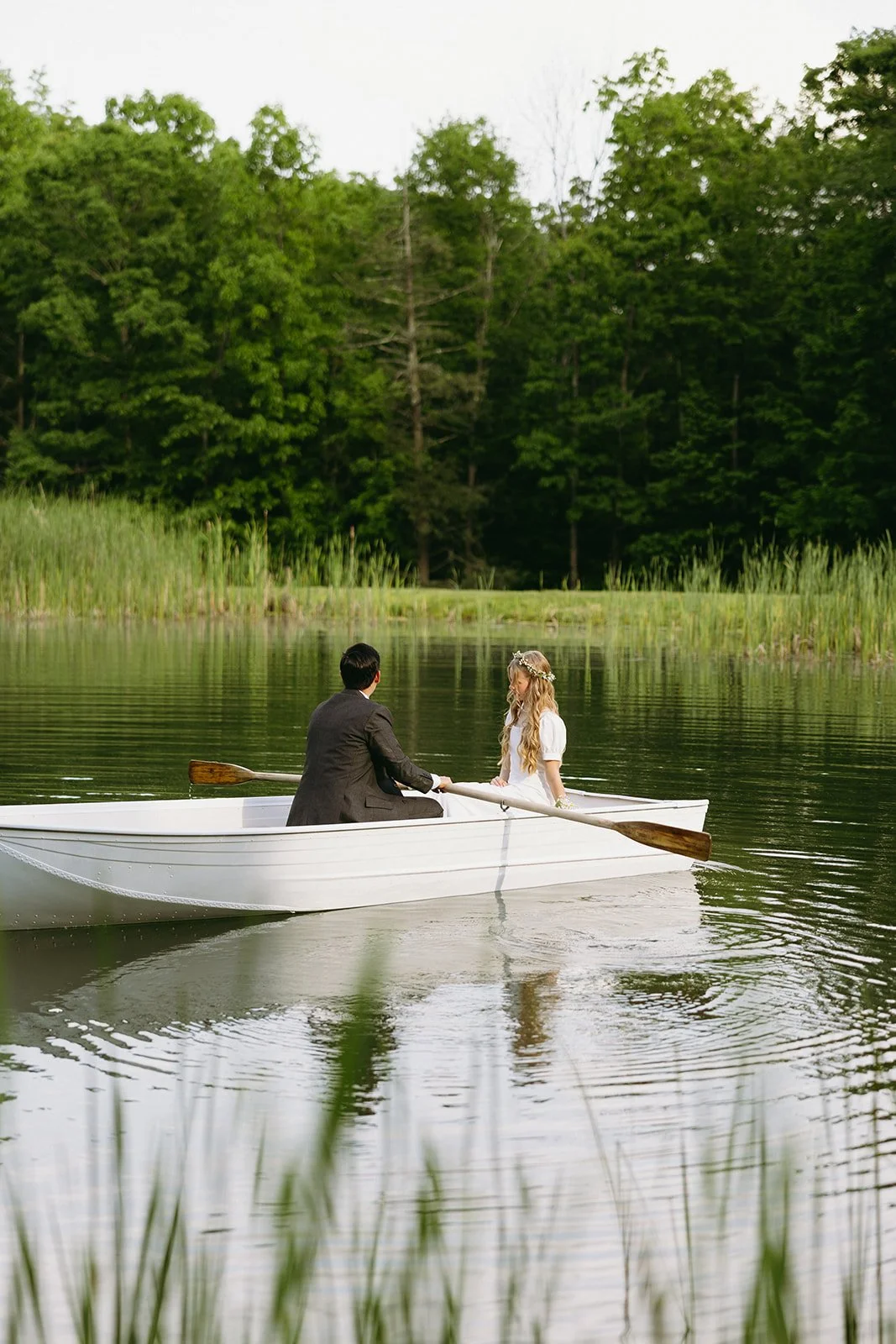 Two people sit and row a small white boat on a calm lake surrounded by tall grass and trees on a summer day for a christ-centered marriage
