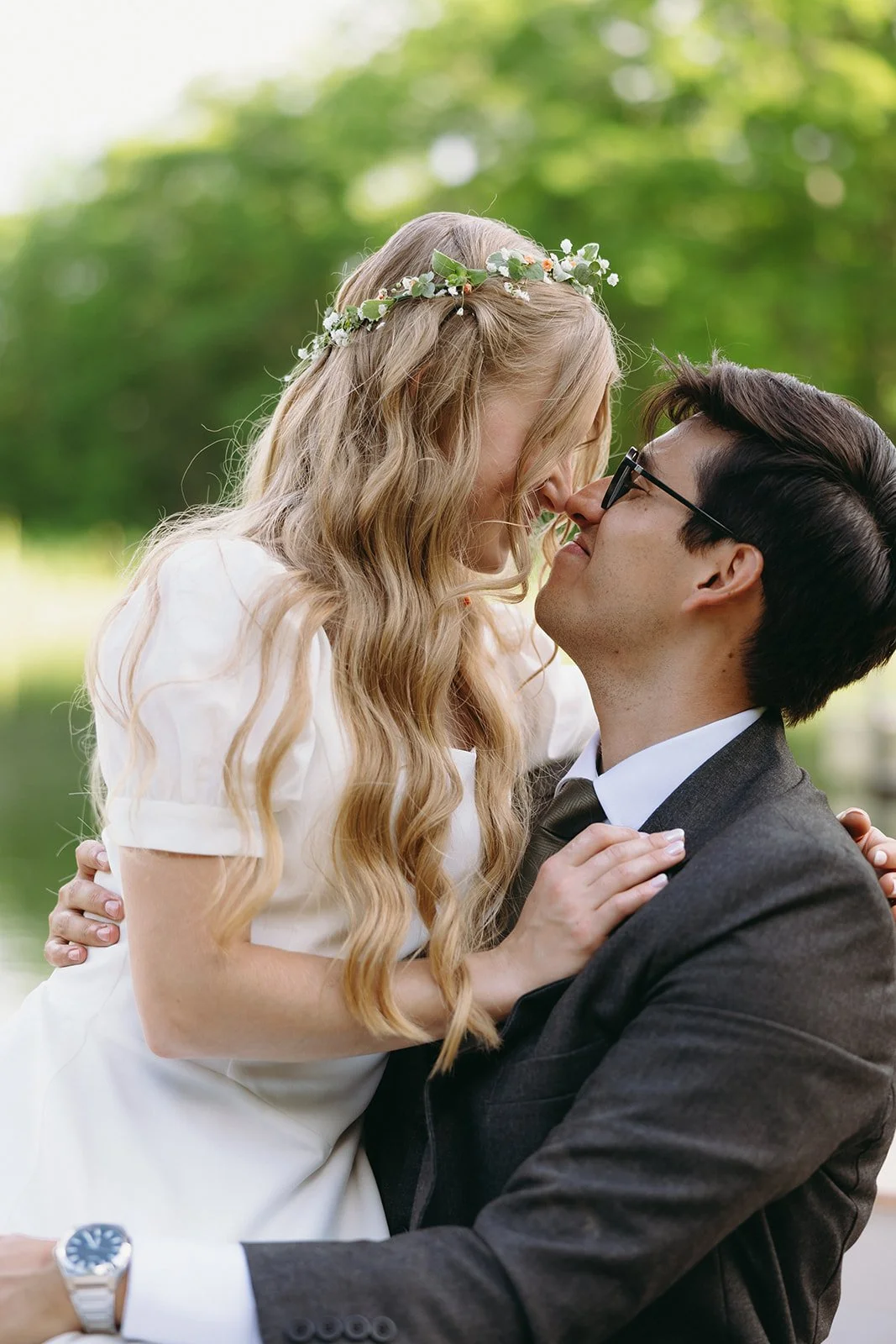 A couple dressed in wedding attire sit closely together outdoors, smiling and touching noses, surrounded by greenery for a christ centered marriage
