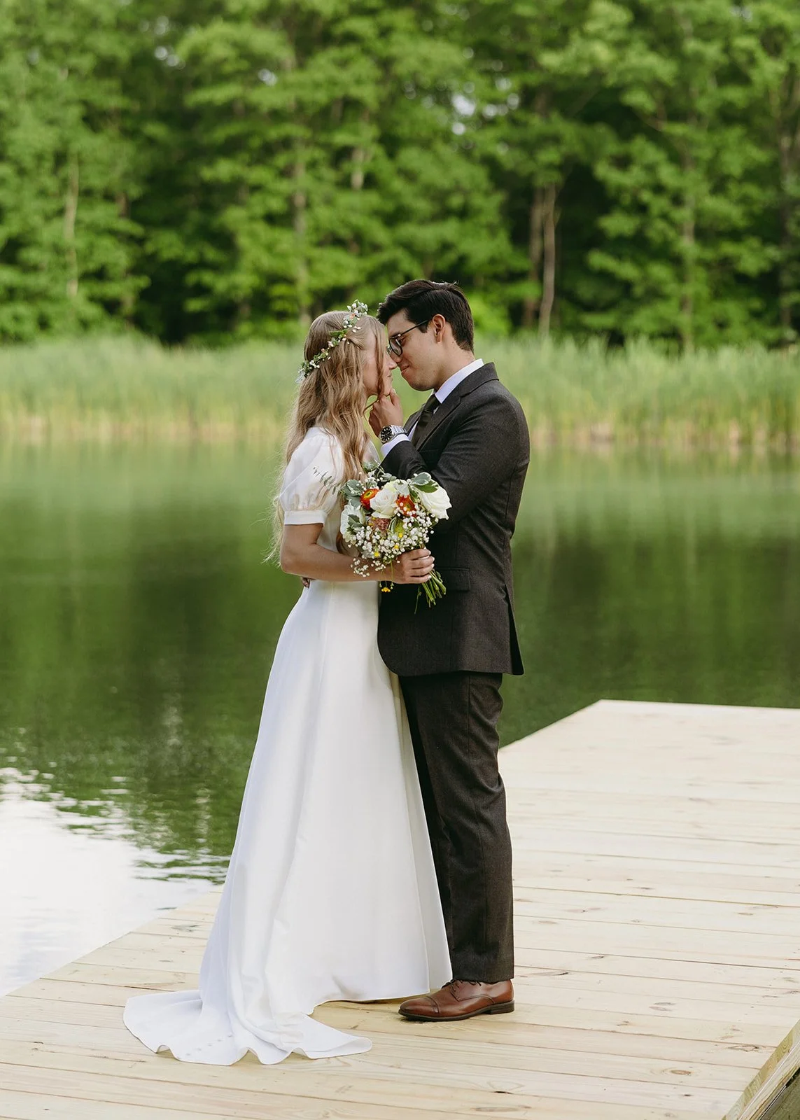 A bride and groom stand on a wooden dock by a lake, embracing and holding a bouquet, with green trees in the background.