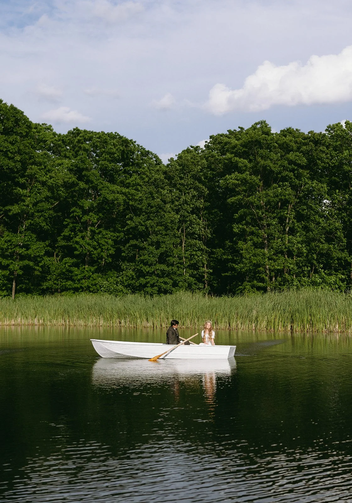 Two people sit in a small white rowboat on a calm lake, surrounded by tall green trees and reeds under a partly cloudy sky for christ-centered marriage