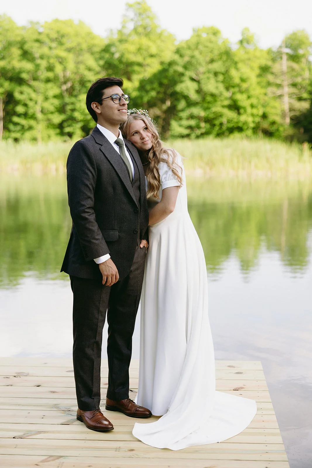 A woman in a white dress leans her head on a man in a dark suit as they stand on a wooden dock by a calm lake, with green trees in the background for a christ-centered marriage 