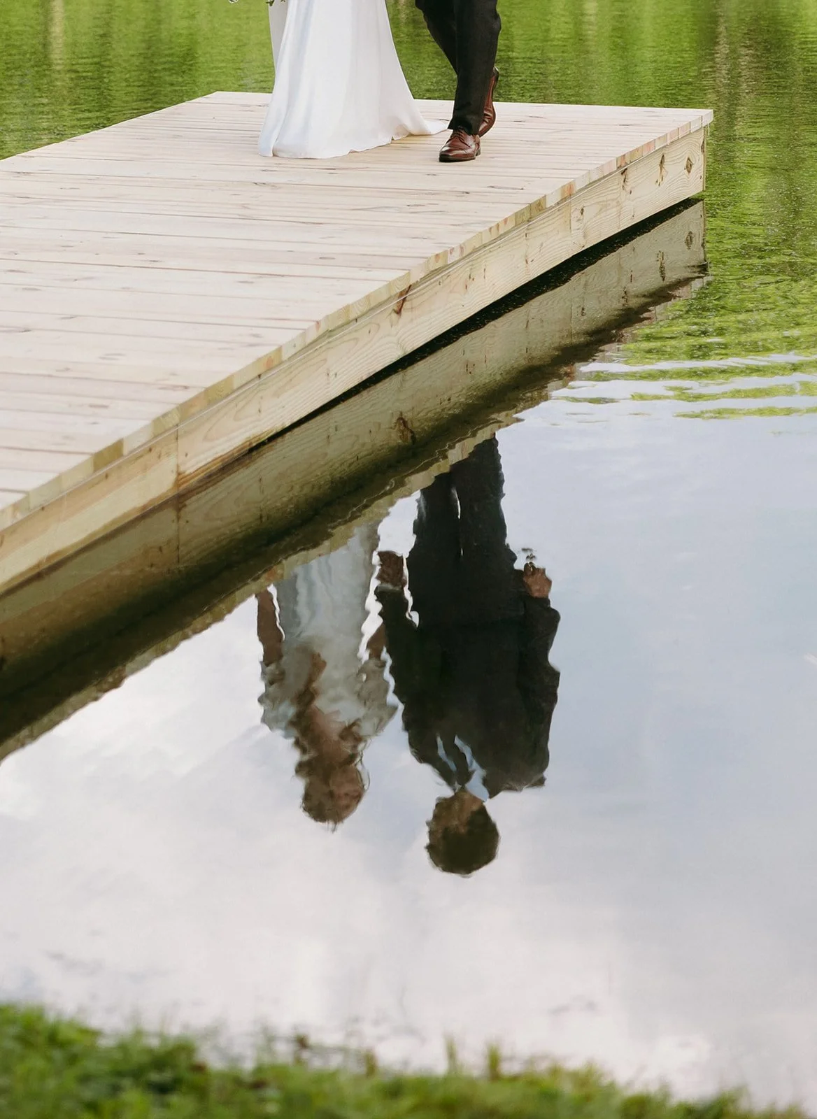 A couple stands on a wooden dock above a pond, their lower bodies and clear reflection visible in the water below.