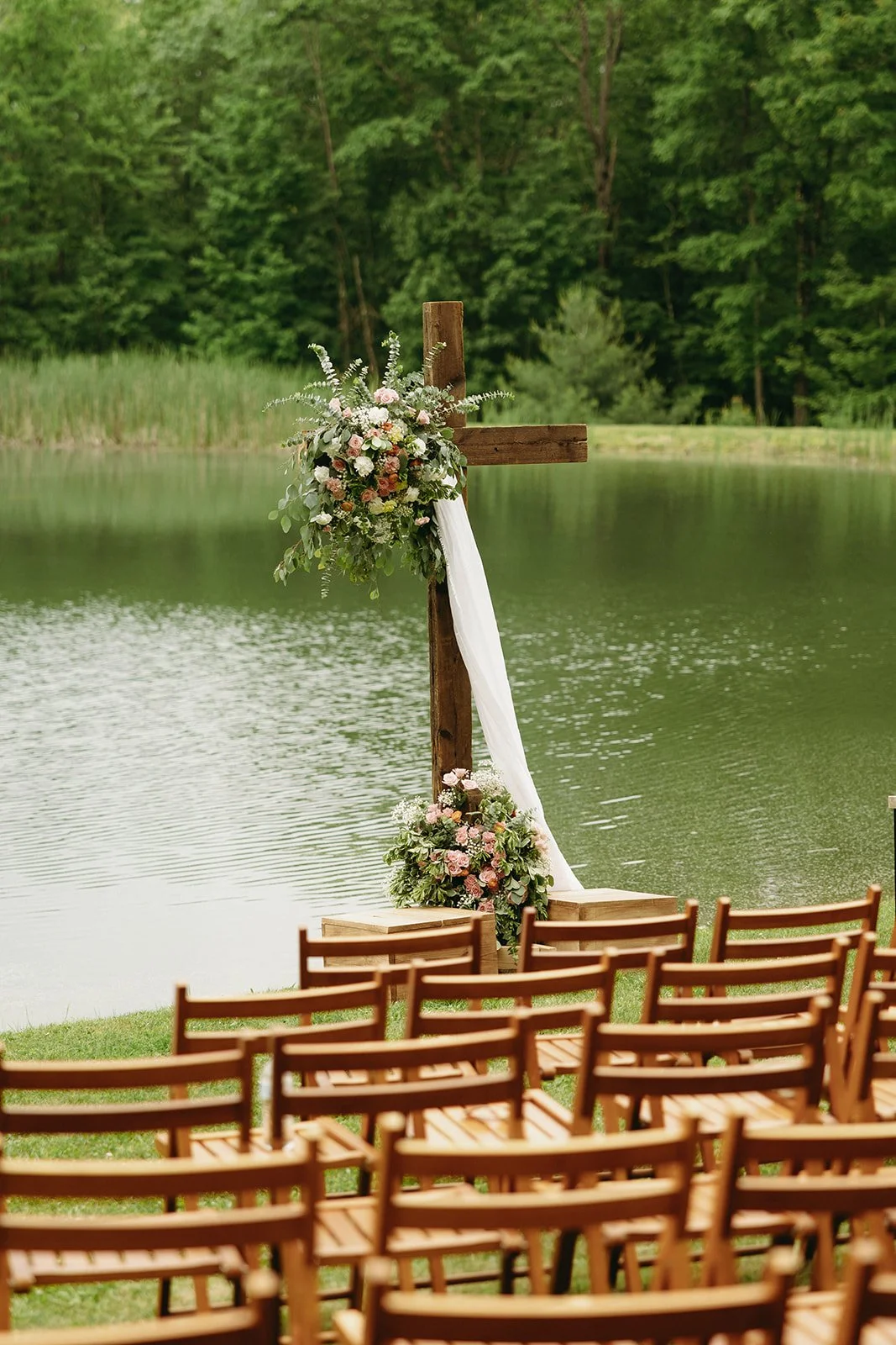 Wooden chairs face a floral-decorated wooden cross by a lake, with lush green trees and grass in the background, set for an outdoor event.