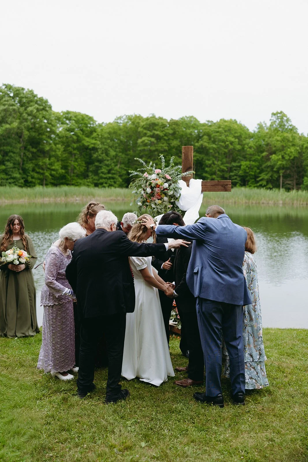 A group of people stand in a circle outdoors near a wooden cross and a pond, hands extended toward a couple in wedding attire.