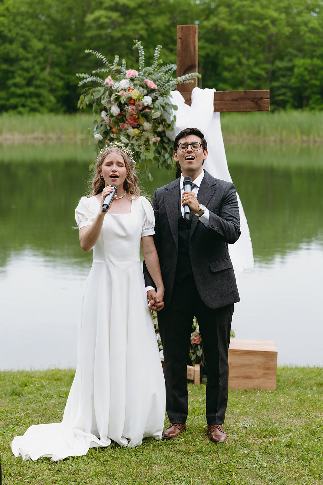 A bride and groom stand outdoors by a lake, holding hands and singing into microphones in front of a flower-adorned wooden cross christ-centered marriage