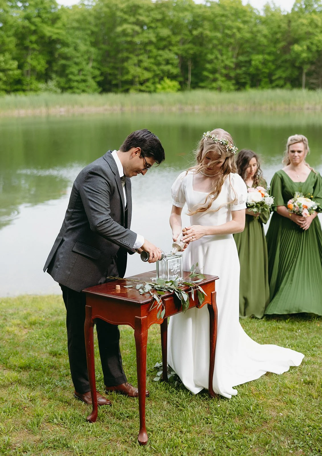 A bride and groom perform a wedding ritual at a small table by a pond, with bridesmaids in green dresses standing nearby.