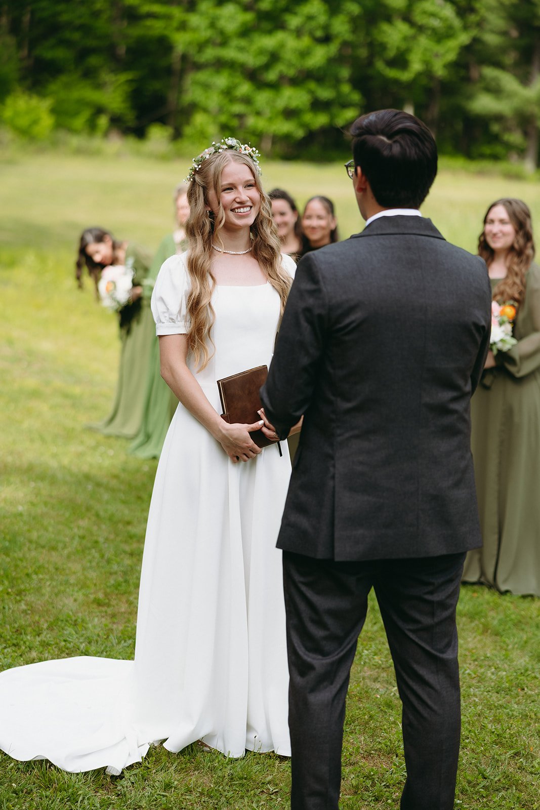 A bride in a white dress and a groom in a suit stand facing each other outdoors, with bridesmaids in green dresses standing behind them.