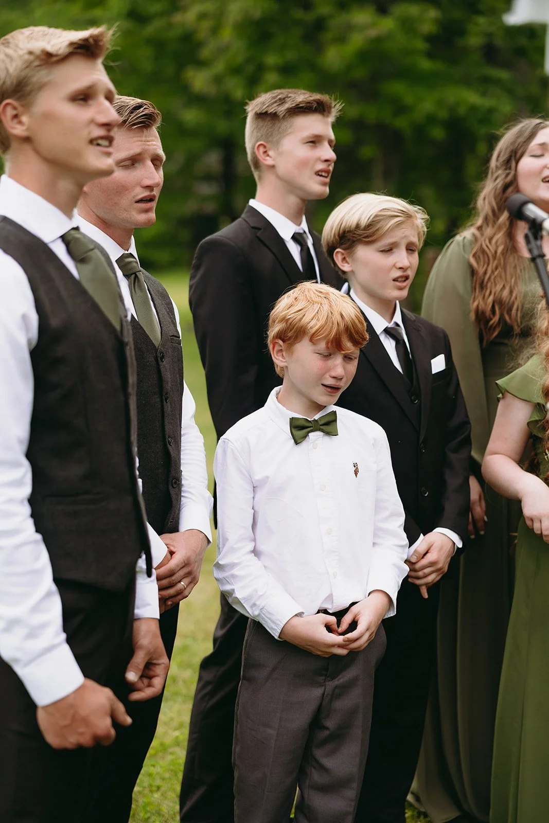 A group of young people dressed formally in black, white, and green outfits sings or poses outdoors on a grassy area.