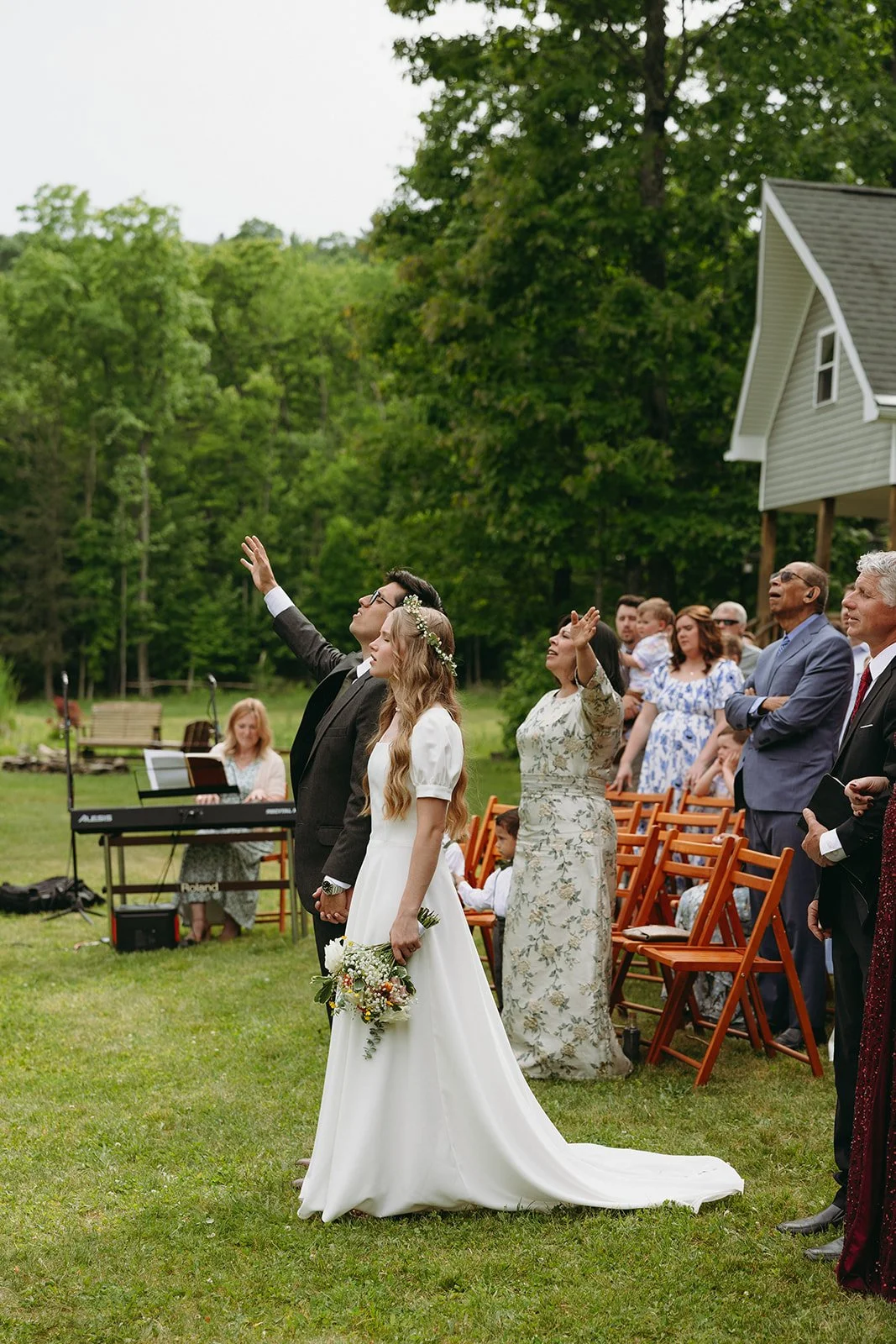 A bride and groom stand outdoors with guests seated behind them, some raising their hands. Trees and a house are in the background for a christ-centered wedding