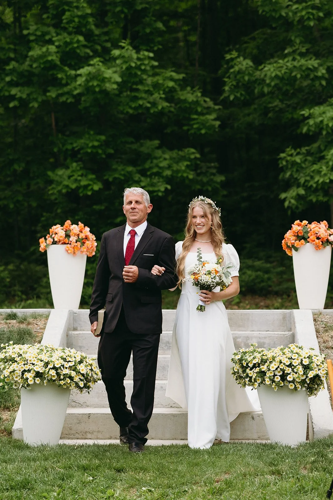 A bride in a white dress and flower crown walks arm in arm with an older man in a suit down outdoor steps lined with flowers.