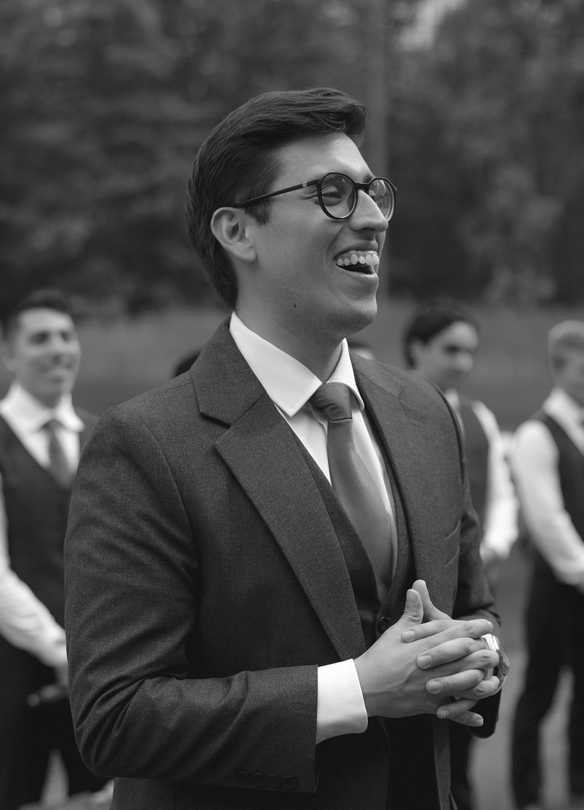 A man in a suit and tie stands outdoors, smiling and clasping his hands, with several people in vests and ties blurred in the background.