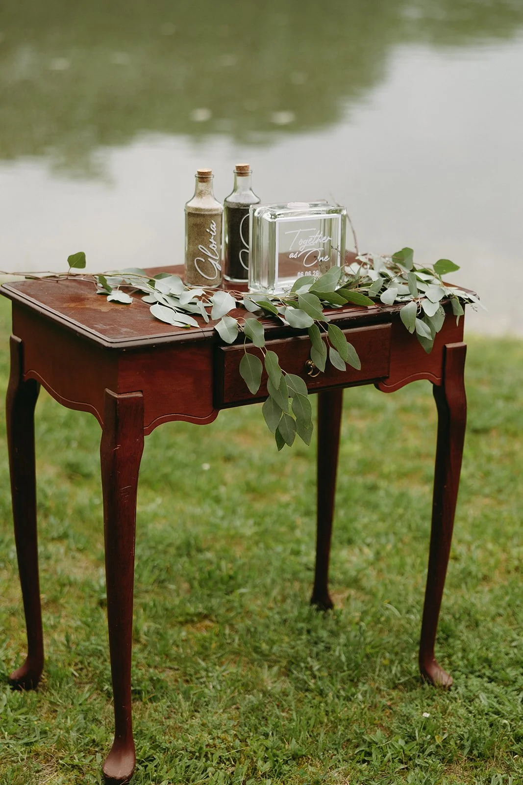 A small wooden table with two bottles, a clear box, and green leafy branches, set on grass near a body of water.