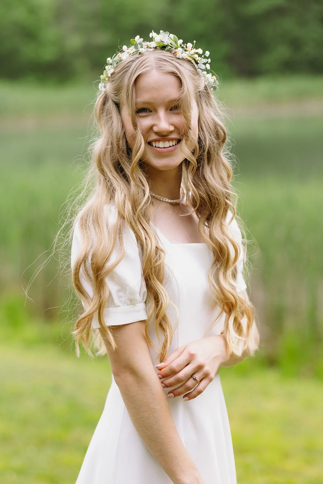 Woman with long wavy blonde hair, wearing a white dress and a floral crown, stands outdoors in front of greenery and smiles at the camera.