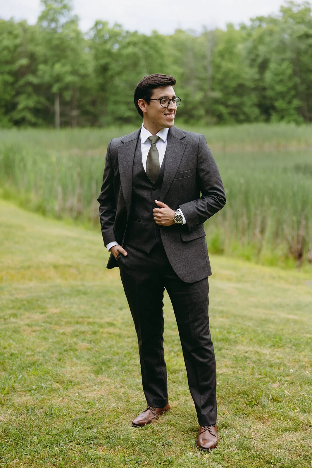Man in a dark suit, white shirt, and tie stands outdoors on grass with trees and tall plants in the background.