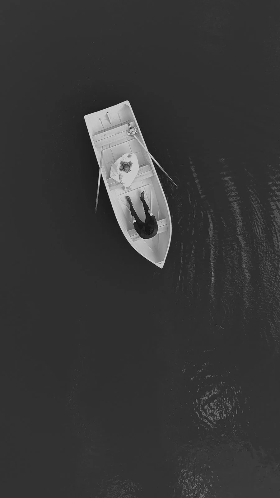 Aerial view of two people sitting in a rowboat on dark water, with one person holding oars and the other facing them for a backyard wedding