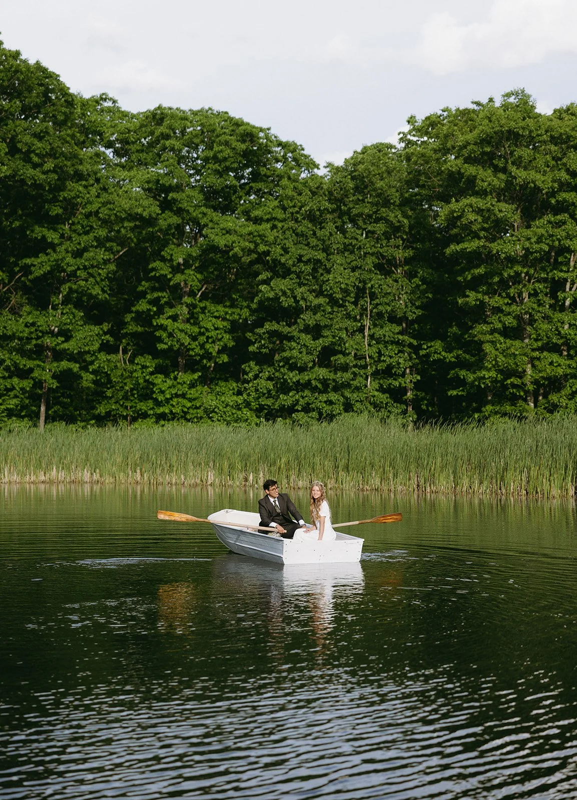 A couple sits together in a white rowboat on a calm lake, surrounded by tall grass and dense green trees under a partly cloudy sky christ-centered wedding