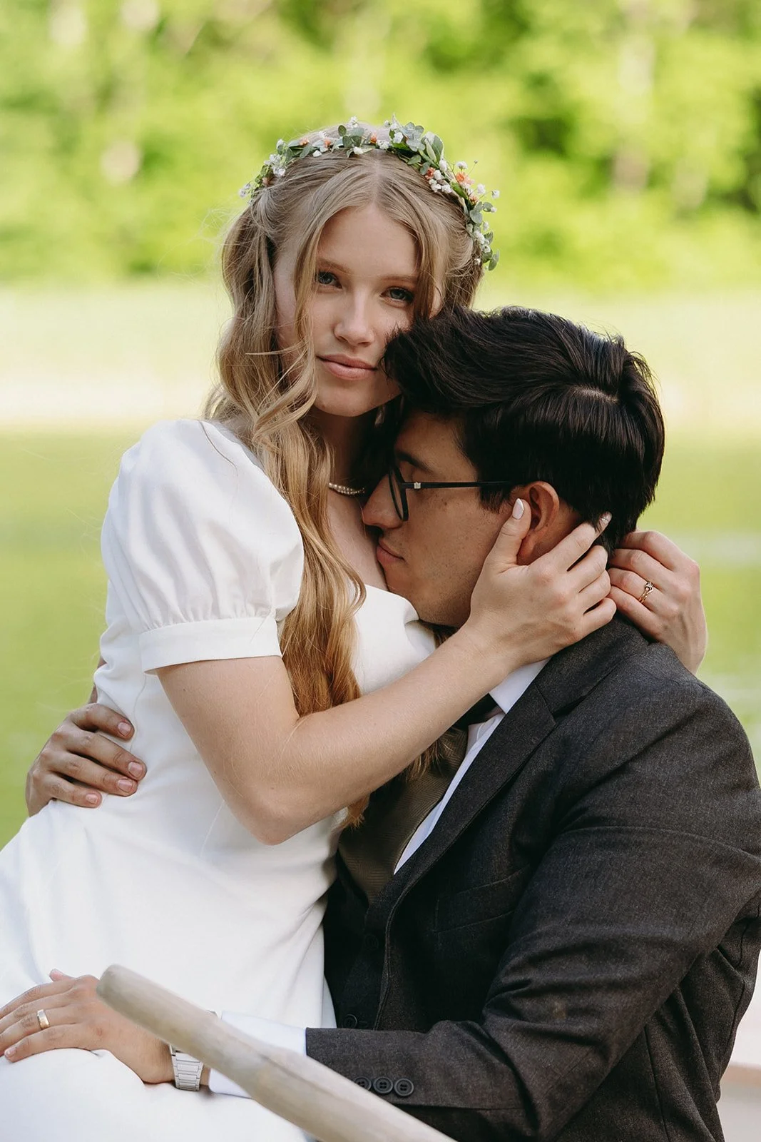 A woman in a white dress with a flower crown embraces a man in a dark suit, sitting outdoors with greenery in the background.