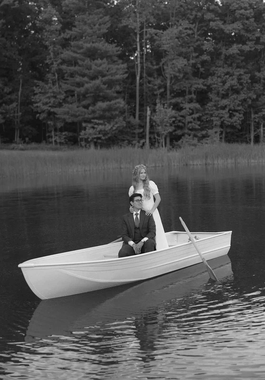 A man in a suit sits in a rowboat on a lake while a woman in a dress stands behind him; trees line the shore in the background for a christ-centered wedding