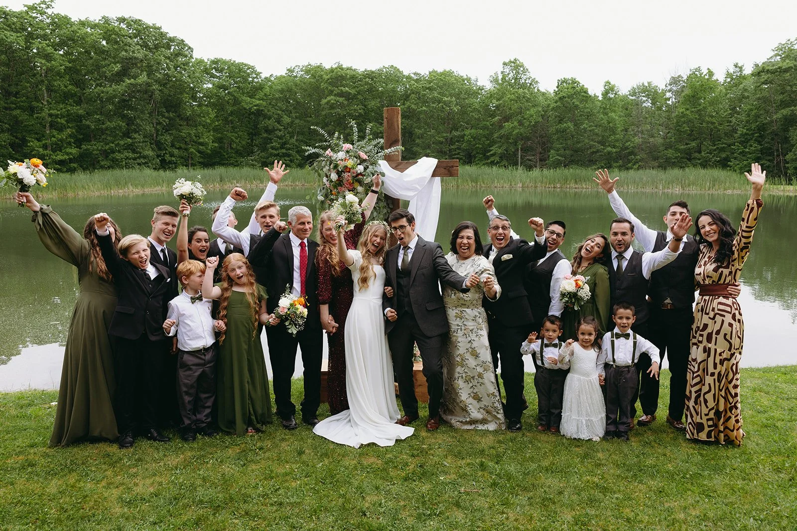 A wedding party poses outdoors by a pond, with the bride and groom in the center, surrounded by family and friends, all smiling and raising their arms for a backyard wedding 