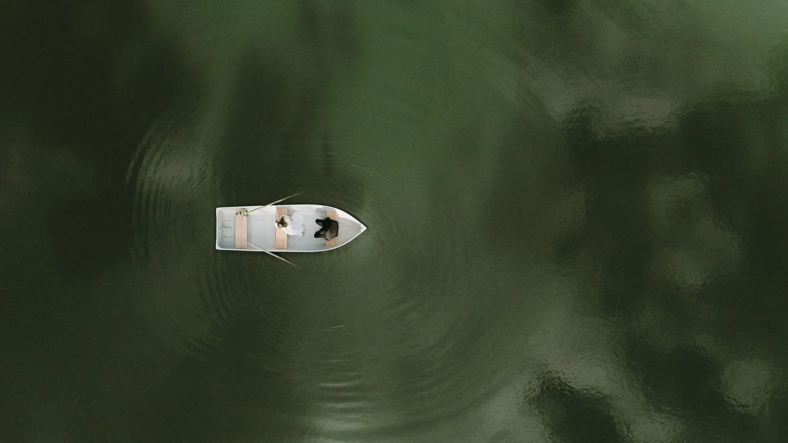 Aerial view of a small white rowboat with one person inside, floating on calm, dark green water.