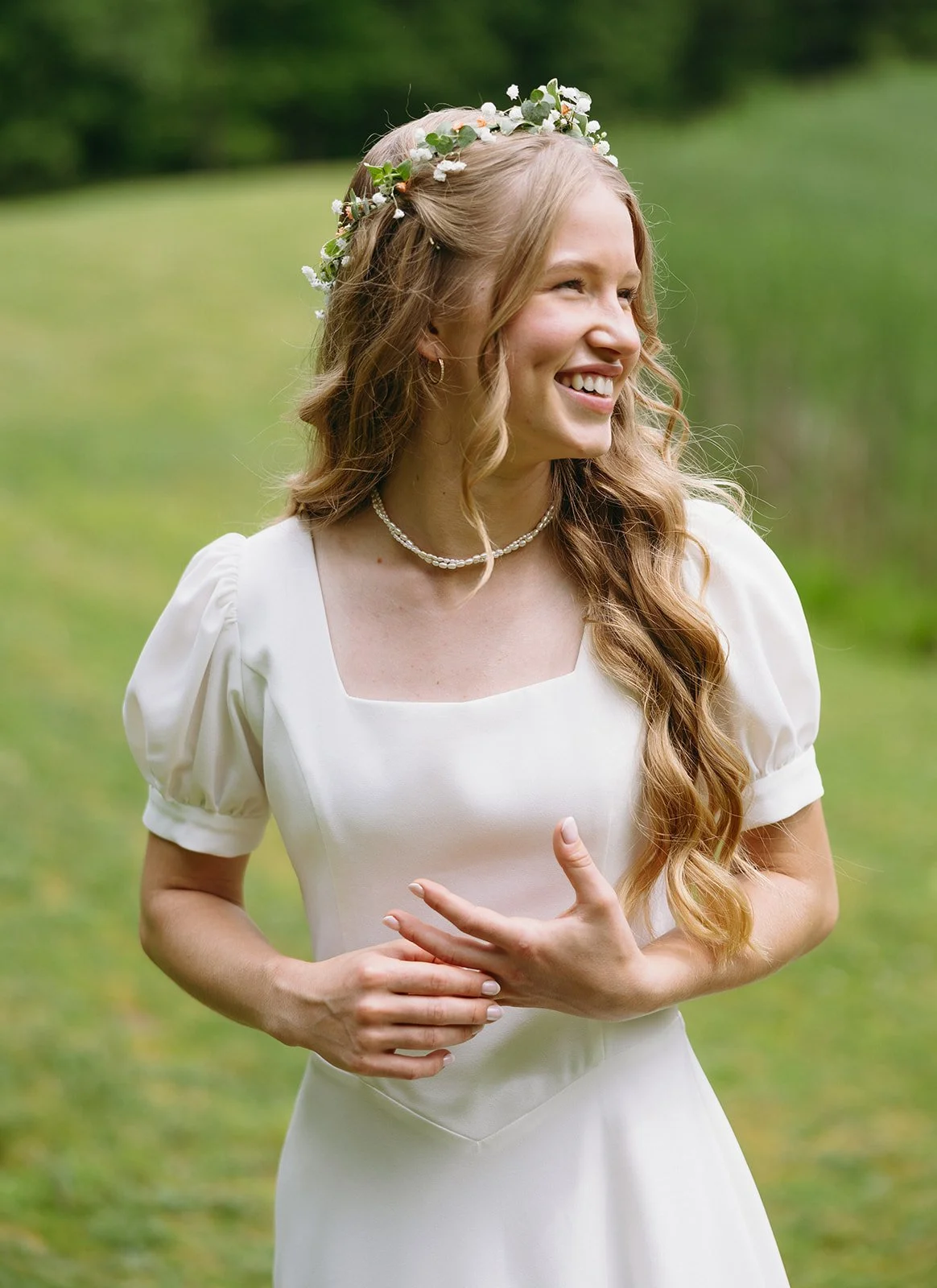 Woman in a white dress with puff sleeves and a flower crown smiles while standing outdoors on green grass for a backyard wedding